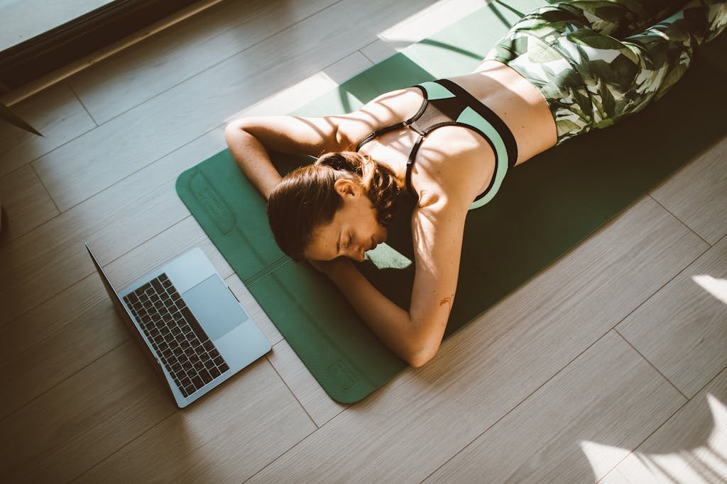 A woman in activewear lies on a yoga mat next to a laptop, relaxing indoors.