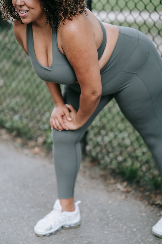 Curvy woman in sportswear stretches in front of a park fence, preparing for exercise.