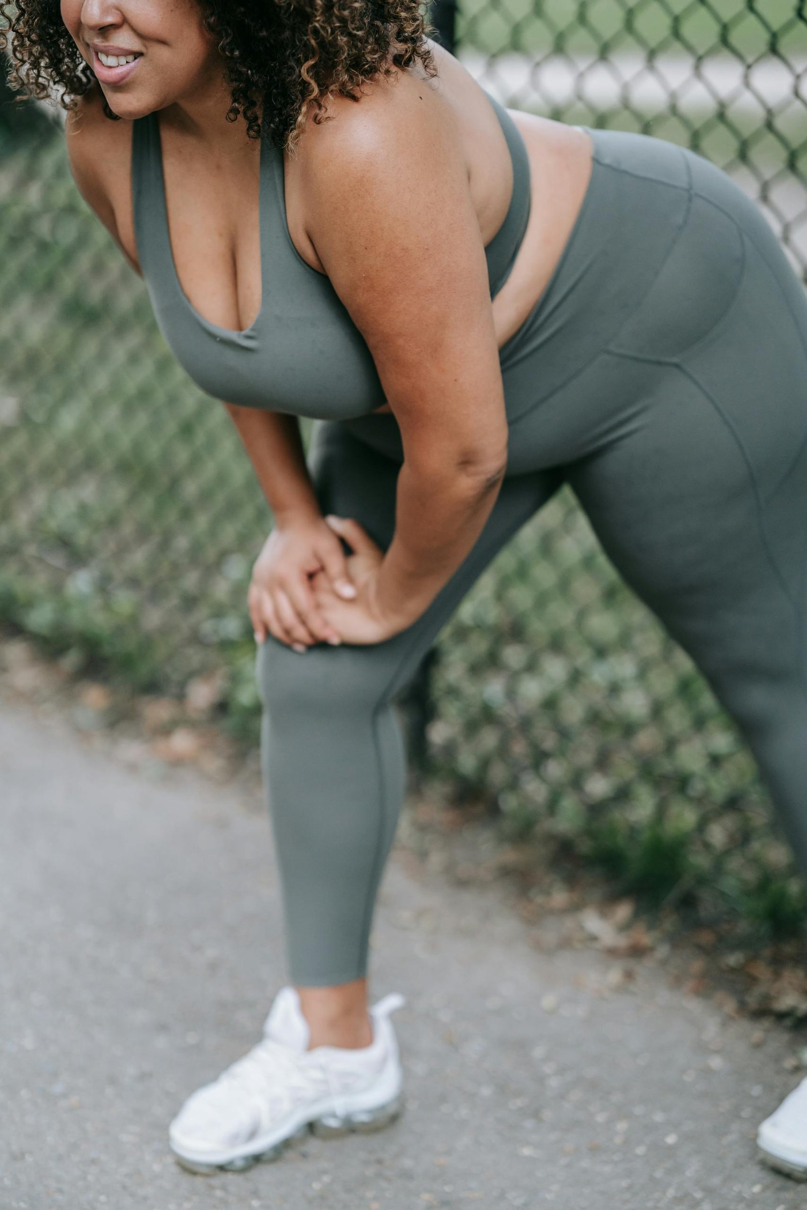 Curvy woman in sportswear stretches in front of a park fence, preparing for exercise.