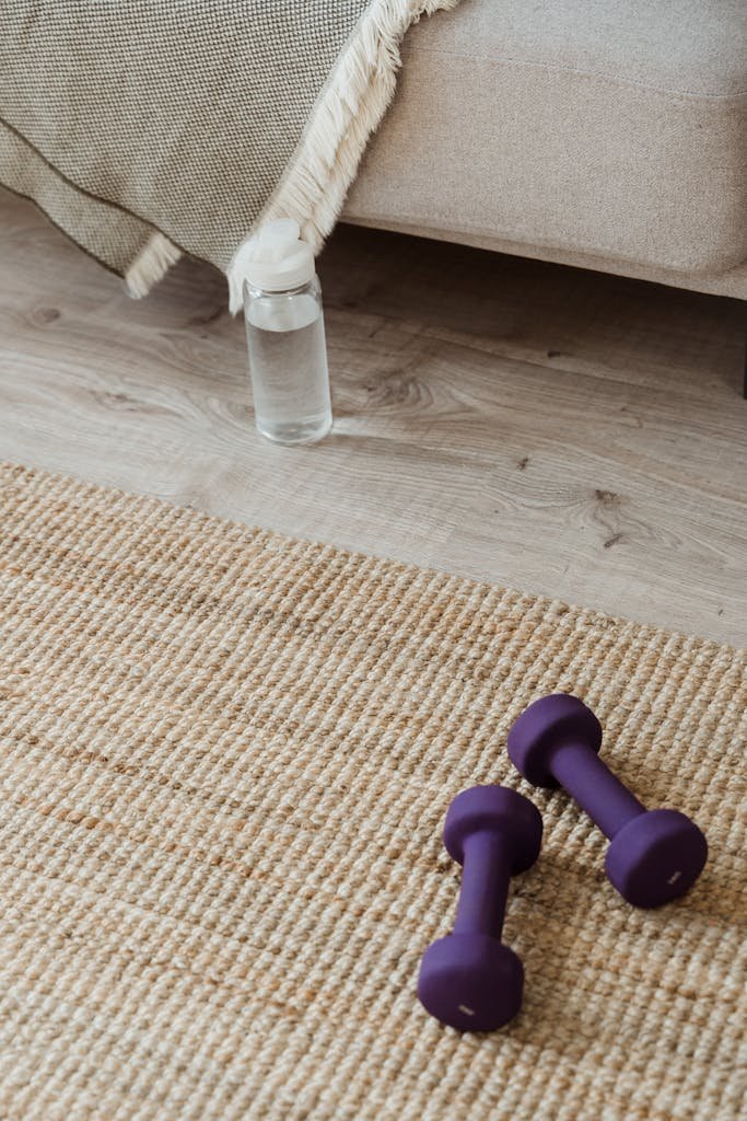 Home workout setup featuring dumbbells, water bottle, and woven mat on wooden floor.